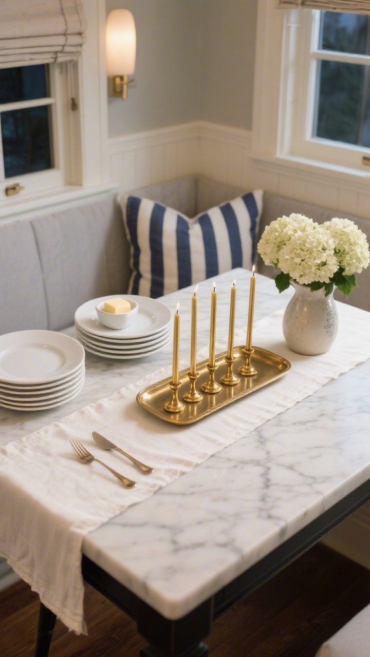 Overhead detail shot of a classic kitchen nook tabletop: honed marble bistro table with an ivory linen runner and a narrow rectangular brass tray centered, holding six slim gold Shabbat tapers in aged gold. Surrounding practical-pretty accents: stacked white plates, a small butter dish, simple flatware, a ceramic pitcher filled with hydrangeas; hints of navy stripe from nearby pillows/roman shade at frame edge. Palette of ivory, soft gray, navy stripe, aged gold. Soft evening glow mingling with gentle ambient light, photorealistic.