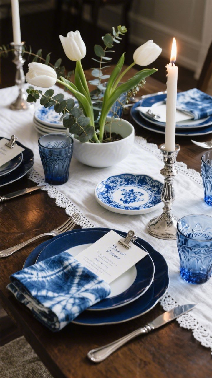 Photorealistic overhead detail shot of an indigo and porcelain heritage table vignette: a crisp white hemstitch runner beneath blue-and-white porcelain plates stacked on navy chargers; brushed-nickel flatware neatly placed. Indigo shibori cloth napkins add pattern, a contemporary white porcelain Seder plate sits nearby, and sleek silver candlesticks frame a low ikebana bowl arranging white tulips and eucalyptus in graceful arcs. Smoky blue water glasses and cut-crystal wine glasses sparkle in candlelight. A small blue calligraphed menu is clipped with a mini silver bulldog clip on the plate. Palette: navy, porcelain white, soft eucalyptus green. Soft evening candlelight; no people.